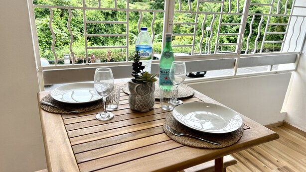 A wooden dining table set with plates, glasses, and a bottle of Pellegrino water, with a view of greenery through a window. - Studio 5 mn des Plages (Schoelcher)