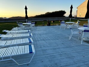 Una terraza de madera con sillones de descanso y mesas blancas, una valla de madera y un edificio al fondo.