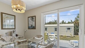 Un coin repas avec une table en verre et des chaises en osier, un lustre et une vue sur une maison, visible à travers une porte-fenêtre coulissante.