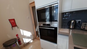 Eine kompakte Küche mit Backofen von Bosch, einem Wasserkocher und einem Toaster. An der Wand hängt eine Rauchmeldeausstattung und ein Mülleimer steht bereit.