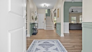 A hallway with a wooden floor, a rug, a staircase, and a living area with a couch. - Pickleball, Arcade & Poker Retreat Near Pinehurst! (Aberdeen)
