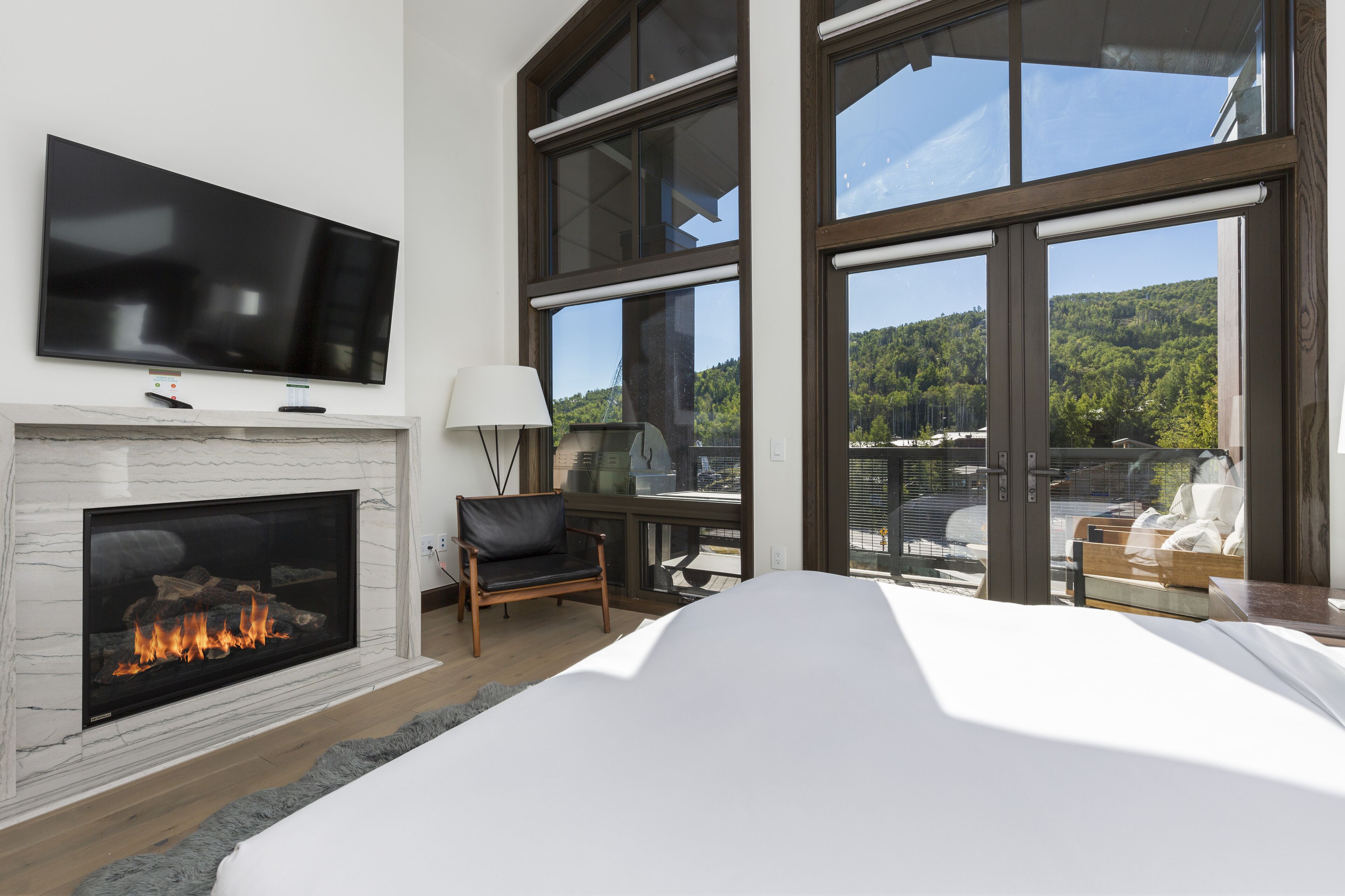 A modern bedroom with a fireplace, a television, and large windows offering a view of a forested area.