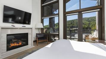 A modern bedroom with a fireplace, a television, and large windows offering a view of a forested area.