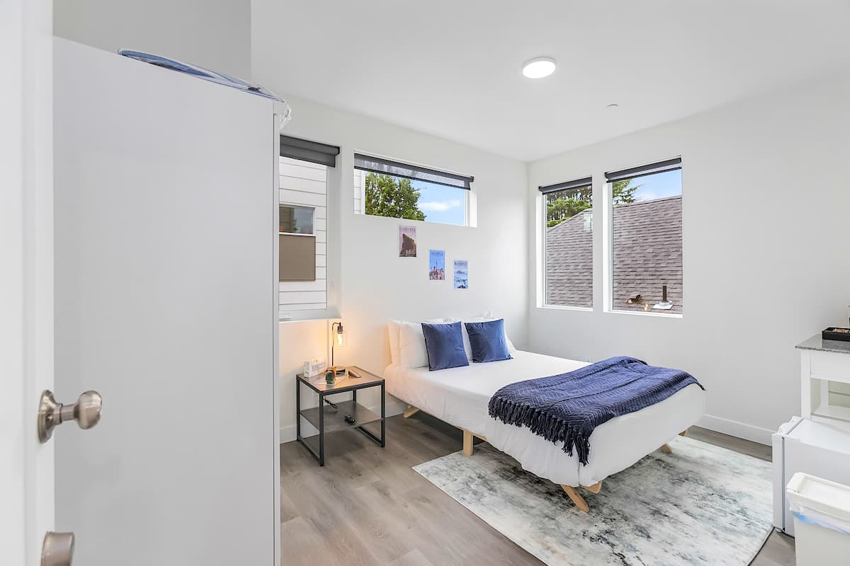 A bedroom with a bed, bedside table, lamp, and two windows.