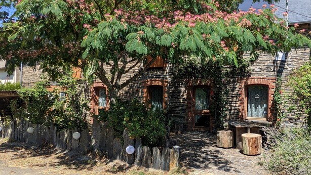 A rustic stone building with arched windows and a tree with pink blossoms in front. - L'HÔT'BERGE de Gannedel (La Chapelle-de-Brain)