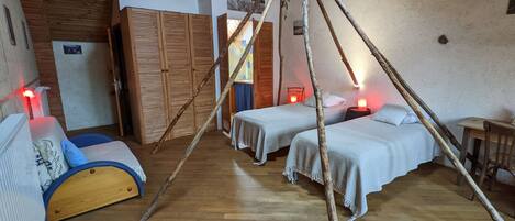 Une chambre rustique avec un plancher en bois, deux lits avec du linge de lit blanc, une armoire en bois et une petite table sur laquelle se trouve une lampe rouge.
