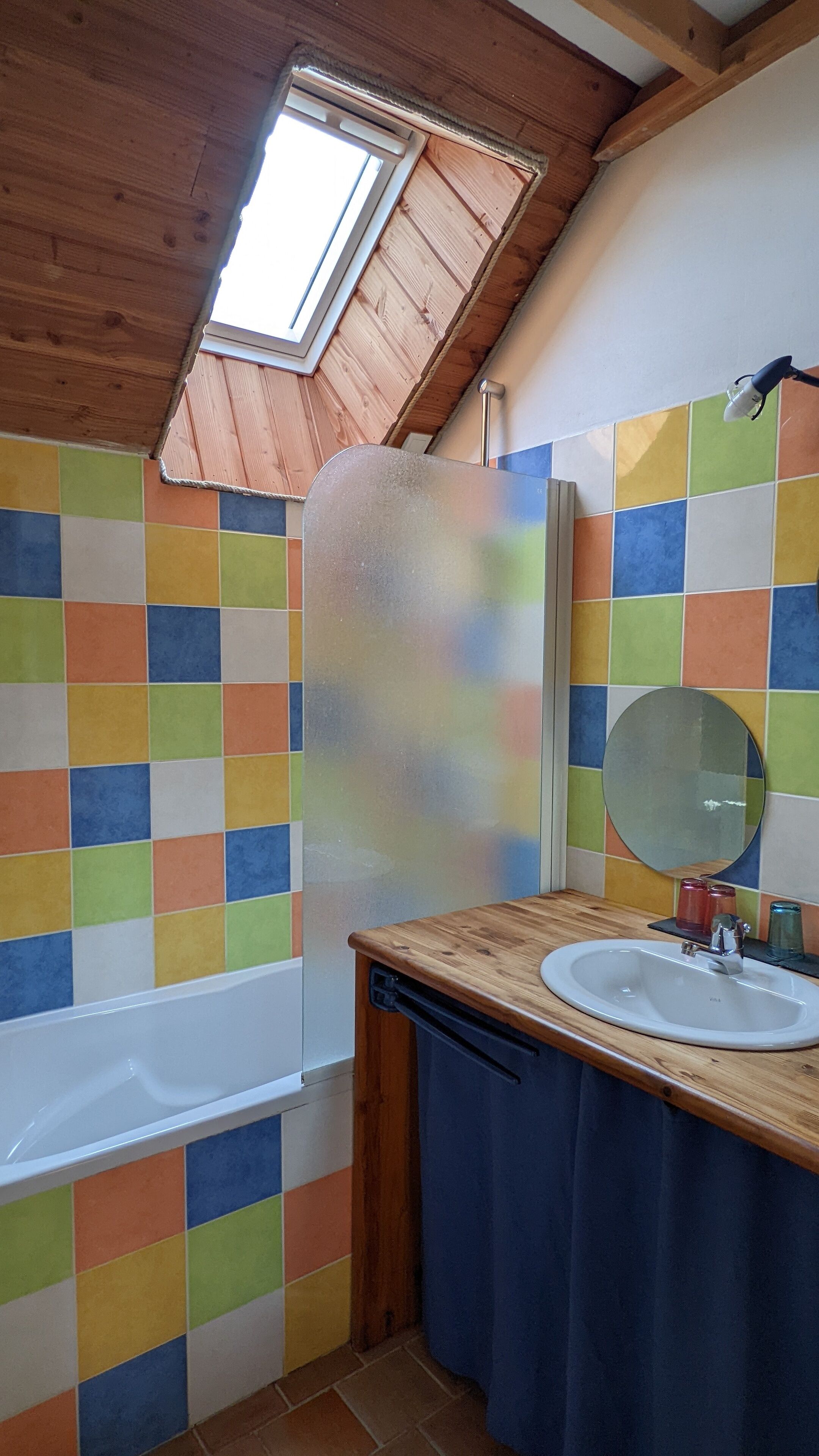 Une salle de bain avec un plafond en bois, un puits de lumière, un mur recouvert de carreaux colorés, un lavabo blanc, un miroir et une baignoire.
