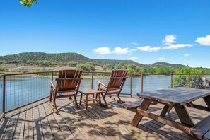 Eine Holzterrasse mit zwei Adirondack-Stühlen und einem Picknicktisch mit Blick auf einen See, vor einer hügeligen Landschaft.