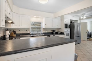 A modern kitchen with white cabinets, a black countertop, stainless steel appliances, and a window with blinds. - Indian Springs | 4BR Lakehouse w/ Fishing & BBQ (Bandera)