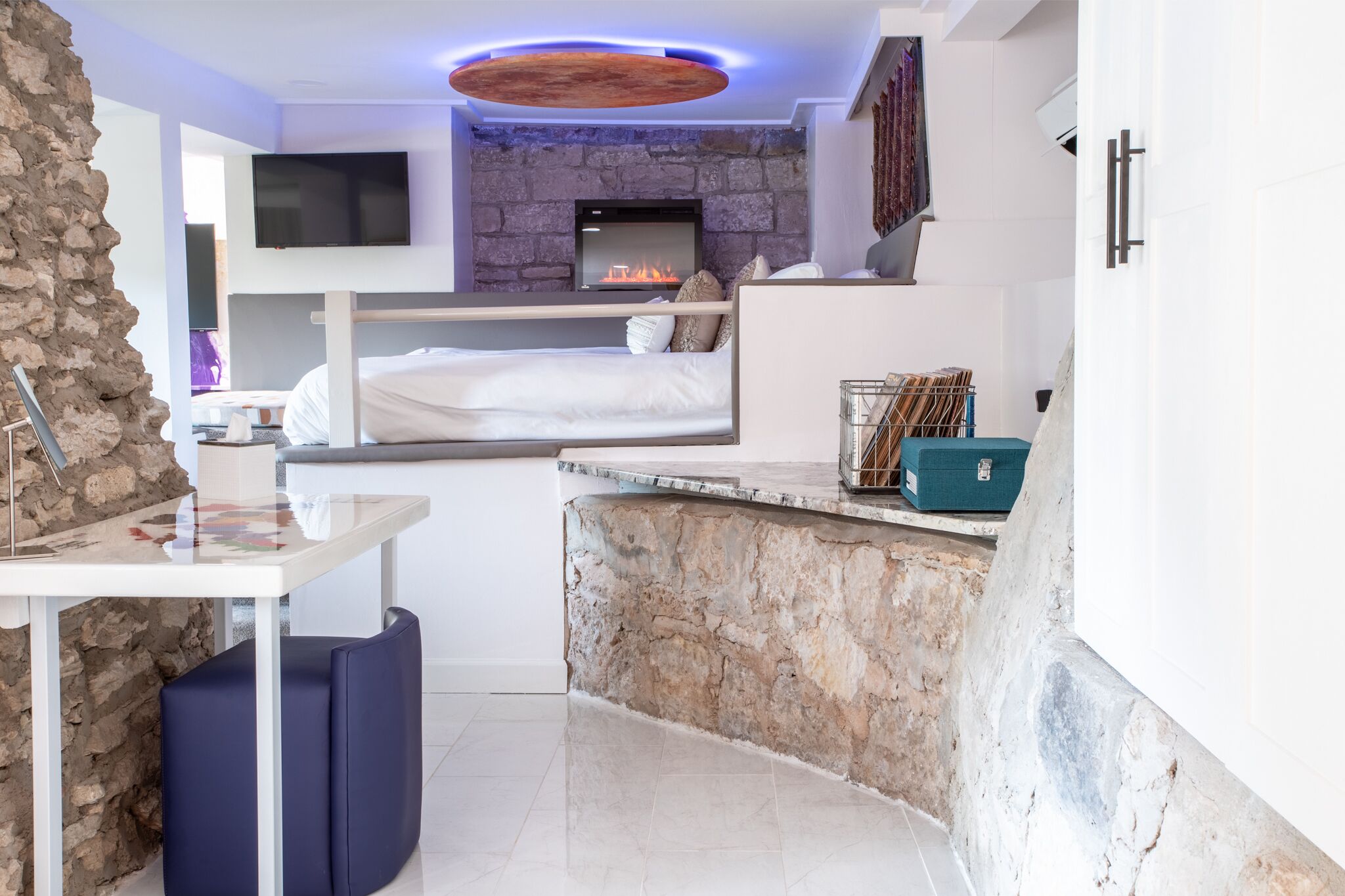 Une chambre moderne avec un mur d’accent en pierre, une cheminée, un lit, une télévision et une petite table avec un ordinateur portable.