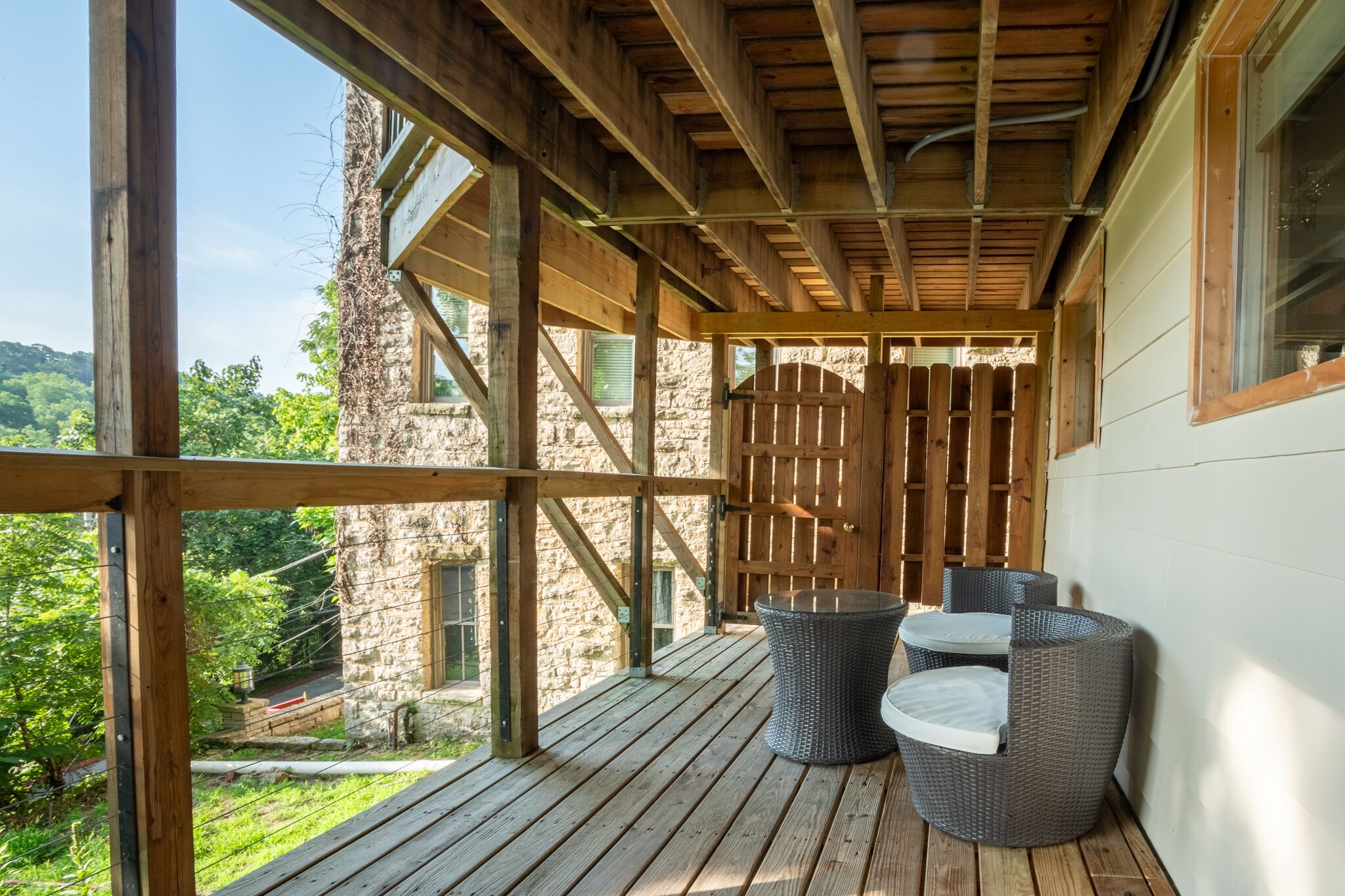 Une terrasse en bois, meublée de meubles en osier, qui offre une vue sur un bâtiment en pierre et une végétation luxuriante.
