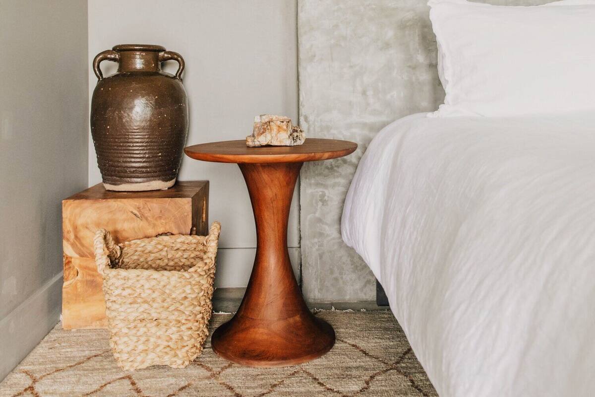 A bedroom with a bed, a hanging light fixture, a wooden side table, a woven basket, and a ceramic vase.