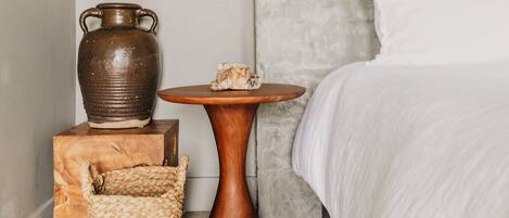 A bedroom with a bed, a hanging light fixture, a wooden side table, a woven basket, and a ceramic vase.