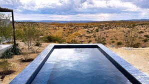 A rectangular swimming pool with blue mosaic tiles, surrounded by desert vegetation, and a view of distant mountains under a cloudy sky. - Willow House (Terlingua)