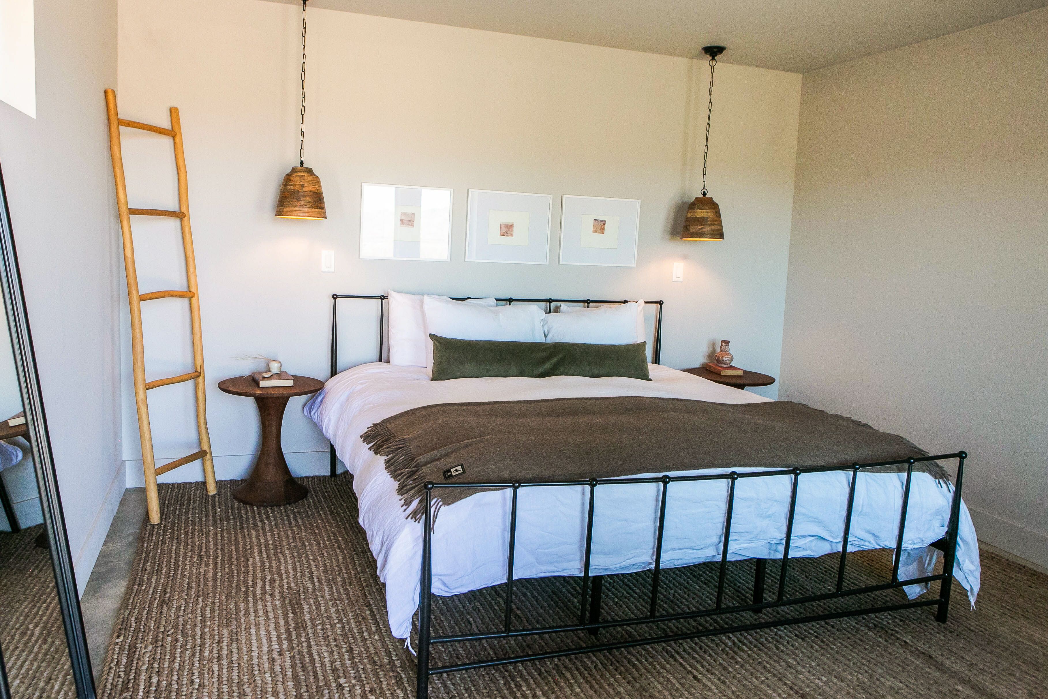 Een keurig opgemaakt slaapkamer met een metalen bed, een houten ladder, een klein rond tafeltje en drie ingekaderde foto’s aan de muur.