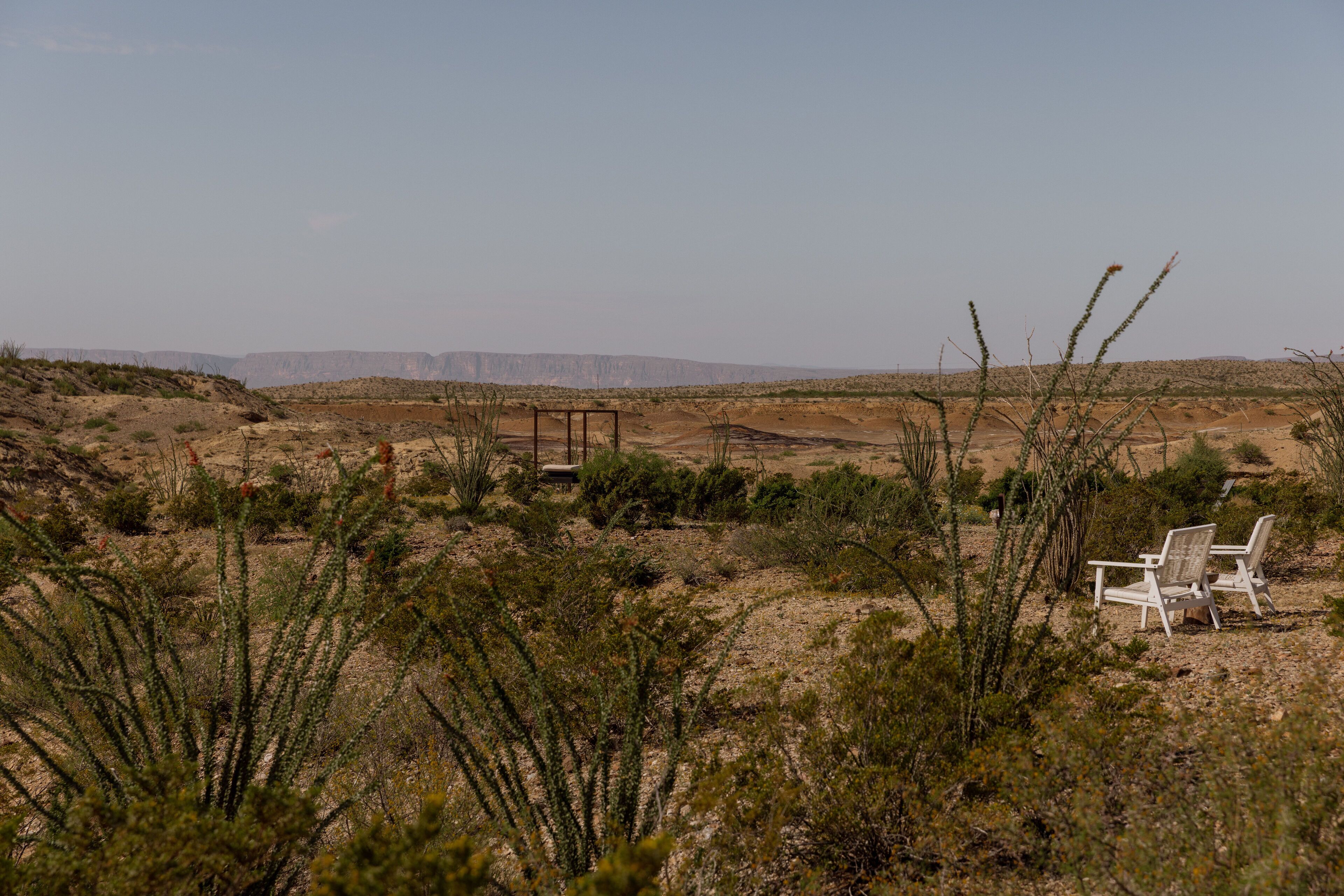 Un paysage désertique avec une végétation clairsemée, une structure en bois et deux chaises blanches.