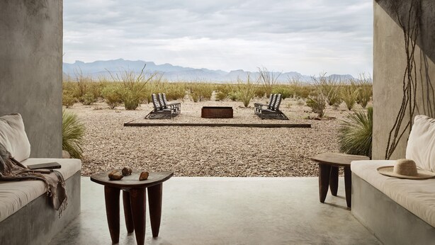 A room with a concrete floor, two sofas, a small table, and two chairs facing a desert landscape with mountains in the distance. - Willow House (Terlingua)