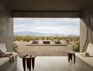 Una habitación con piso de concreto, dos sofás, una mesita y dos sillas mirando un paisaje desértico con montañas al fondo.