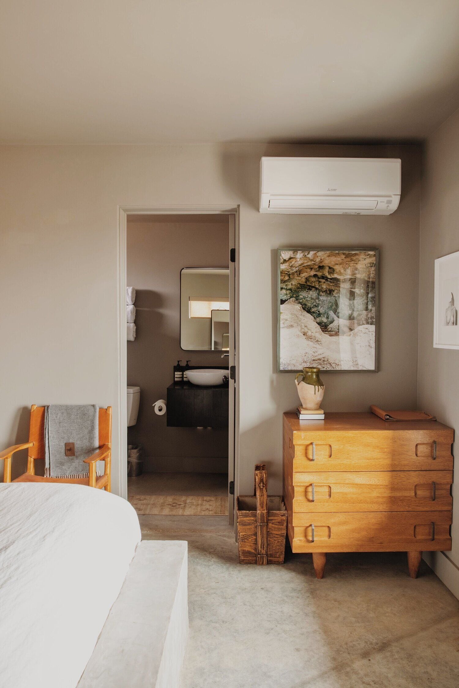 A bedroom with a bed, a wooden dresser, a painting, and a bathroom visible through an open door.