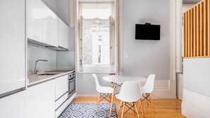 Een moderne keuken met witte kastjes, een ingebouwde oven en een raam met uitzicht op een gebouw. Er staat een witte eettafel met twee stoelen en een vloerkleed met een patroon.