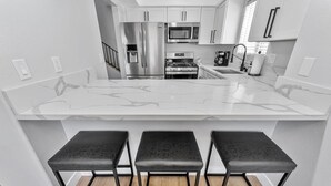 A modern kitchen with white cabinetry, a stainless steel refrigerator, a microwave, a stove, and three black stools with metal frames. - Right By The Sand, Entire 3-Bedroom House In Belmont Shore (Long Beach)