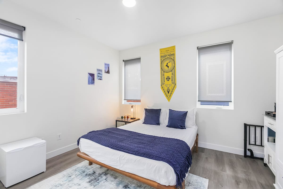 Une chambre à coucher comprenant un lit, une table de chevet, une lampe, un radiateur et une fenêtre avec des stores.