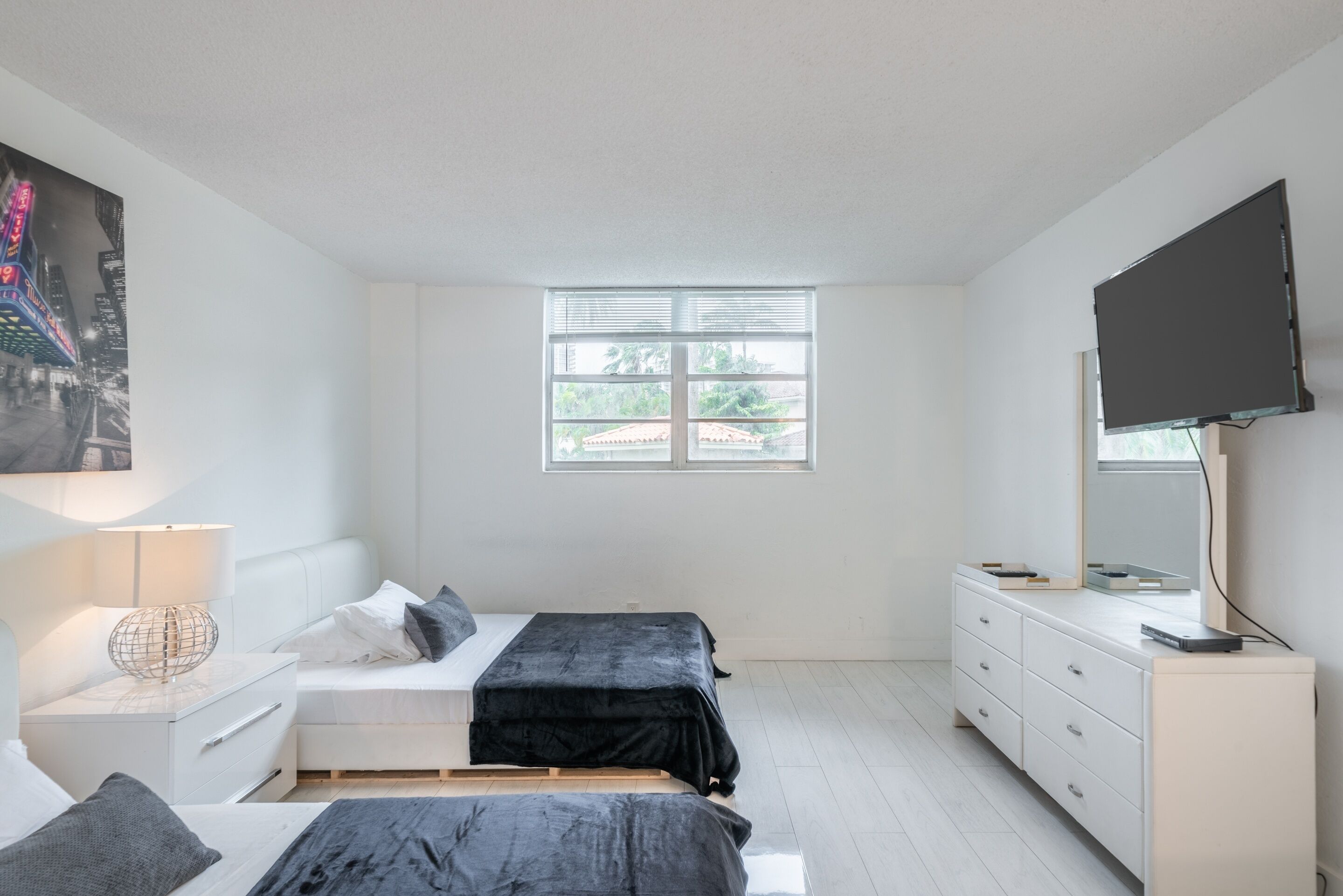 A modern bedroom with two beds, a TV, a dresser, and a window with blinds.