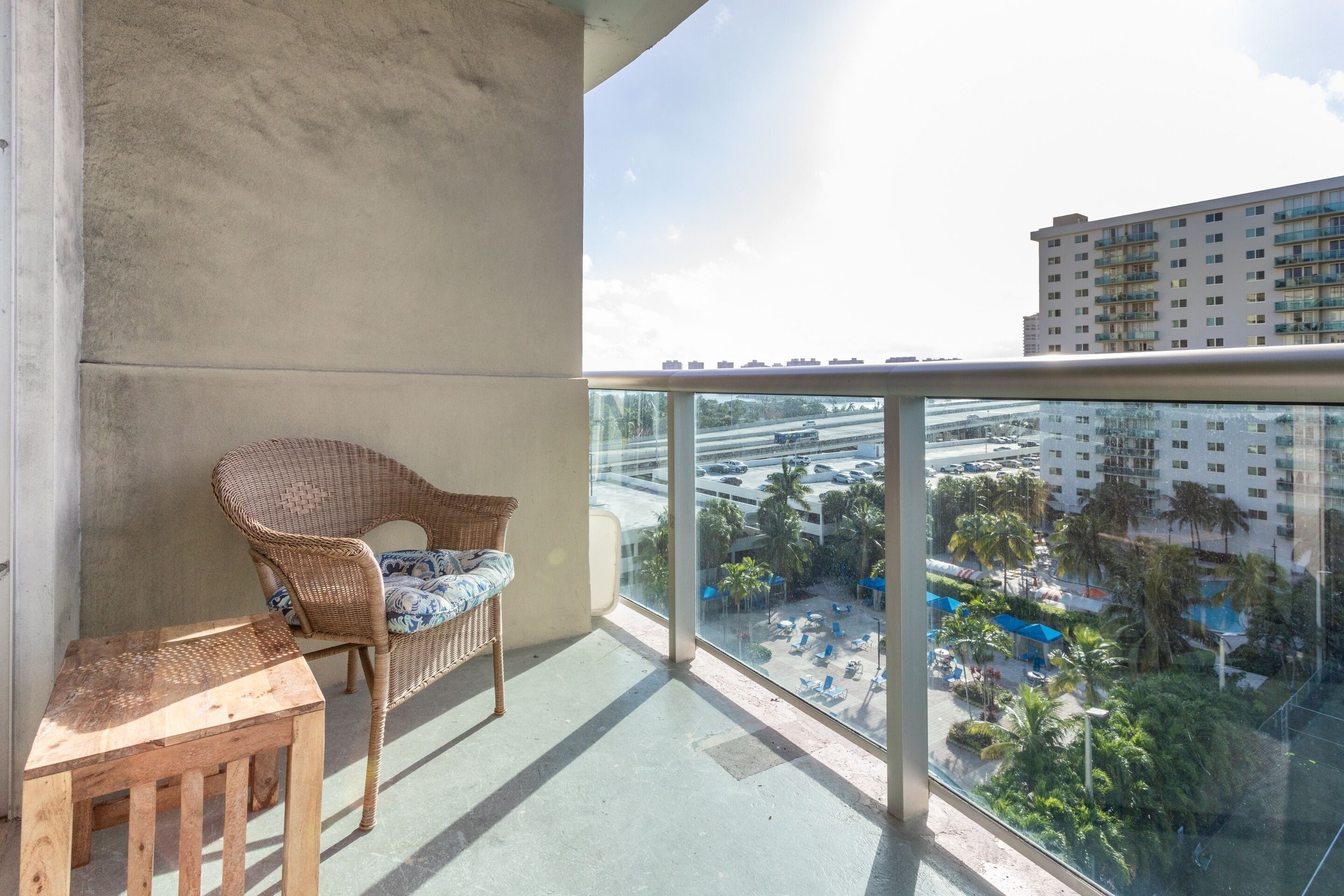 A balcony with a wicker chair and a small wooden table, overlooking a parking area and a building with multiple floors.