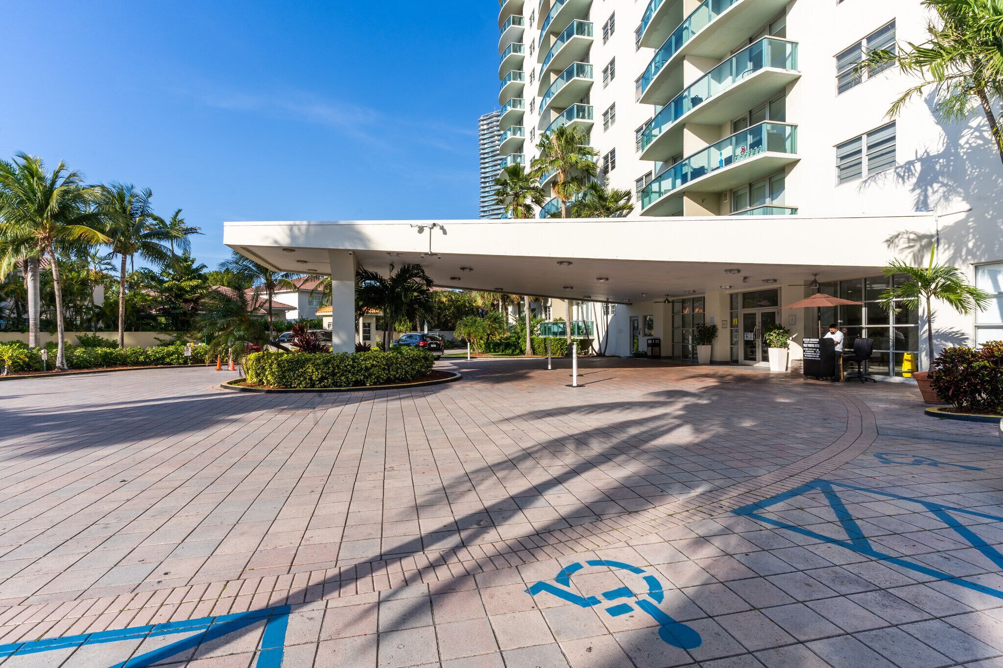 A modern hotel entrance with a covered walkway, palm trees, and a clear blue sky.