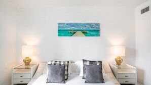 Une chambre à coucher avec un lit, deux tables de chevet avec des lampes, un tableau représentant une jetée et un mur blanc.