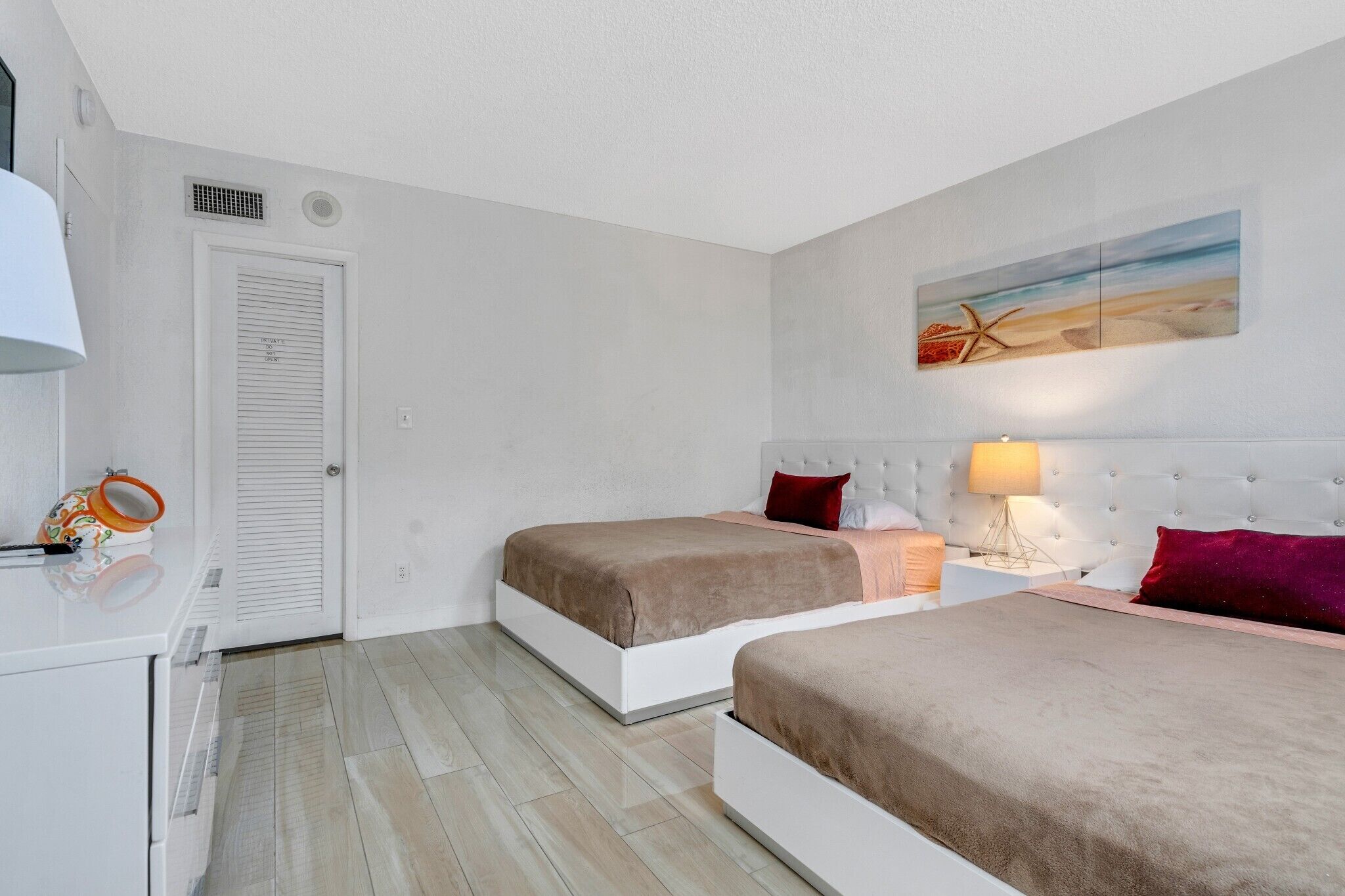 A bedroom with two beds, a white wardrobe, a small table with a lamp, and a beach-themed painting on the wall.