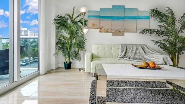 Un salon moderne avec une grande fenêtre, un canapé, une table basse sur laquelle se trouvent des fruits, et un tableau représentant une scène de plage.