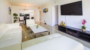 Un salon moderne avec un téléviseur à écran plat, des canapés blancs, une table basse et une cuisine en arrière-plan.