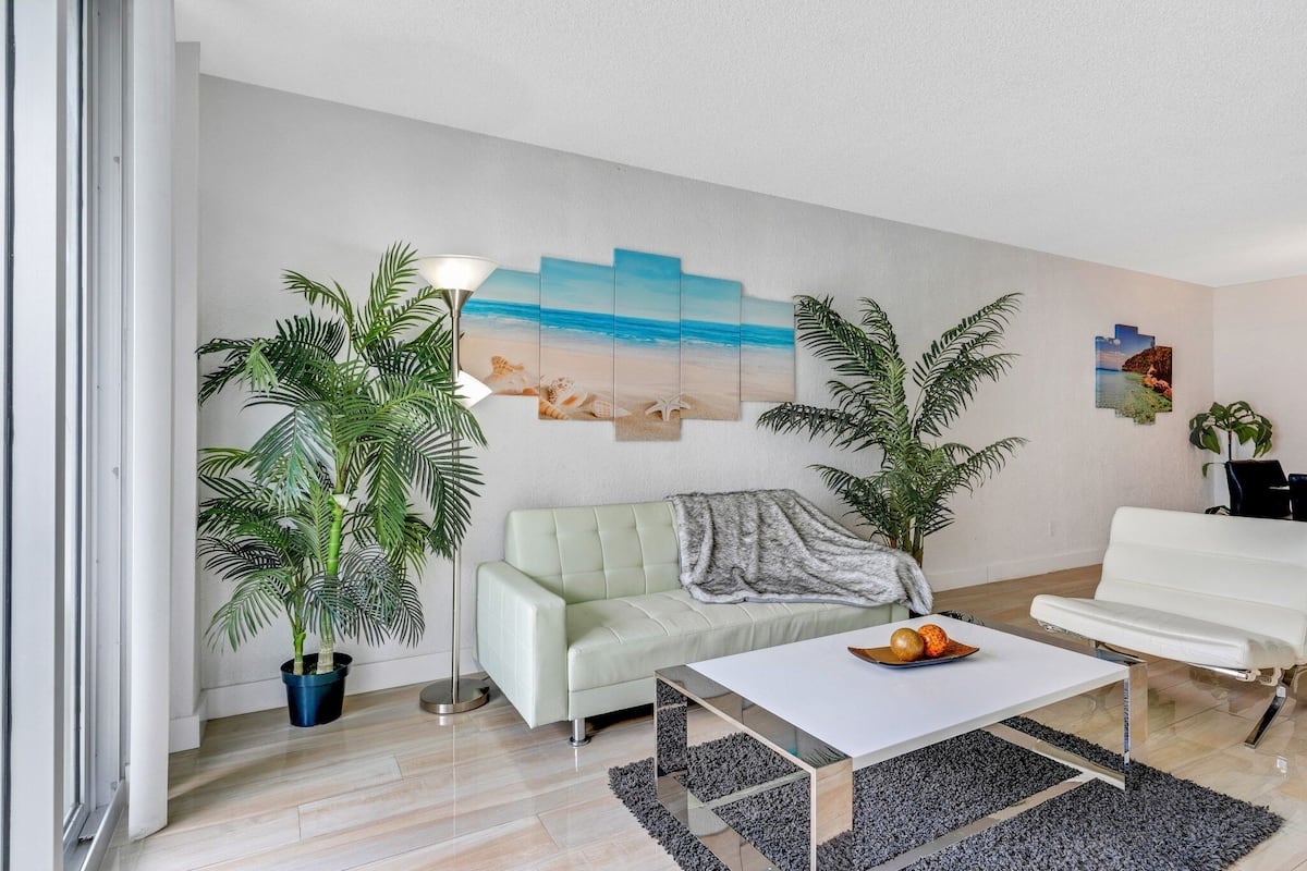 A modern living room with a light green sofa, a white coffee table with fruit, a large painting of a beach scene, and two potted plants.