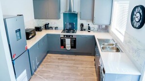 A compact kitchen with grey cabinets, a built-in oven, a stove, a sink, and a clock on the wall. - Cosy 3 Bedroom House Set In Manchester Free wifi & private parking (Greater Manchester)