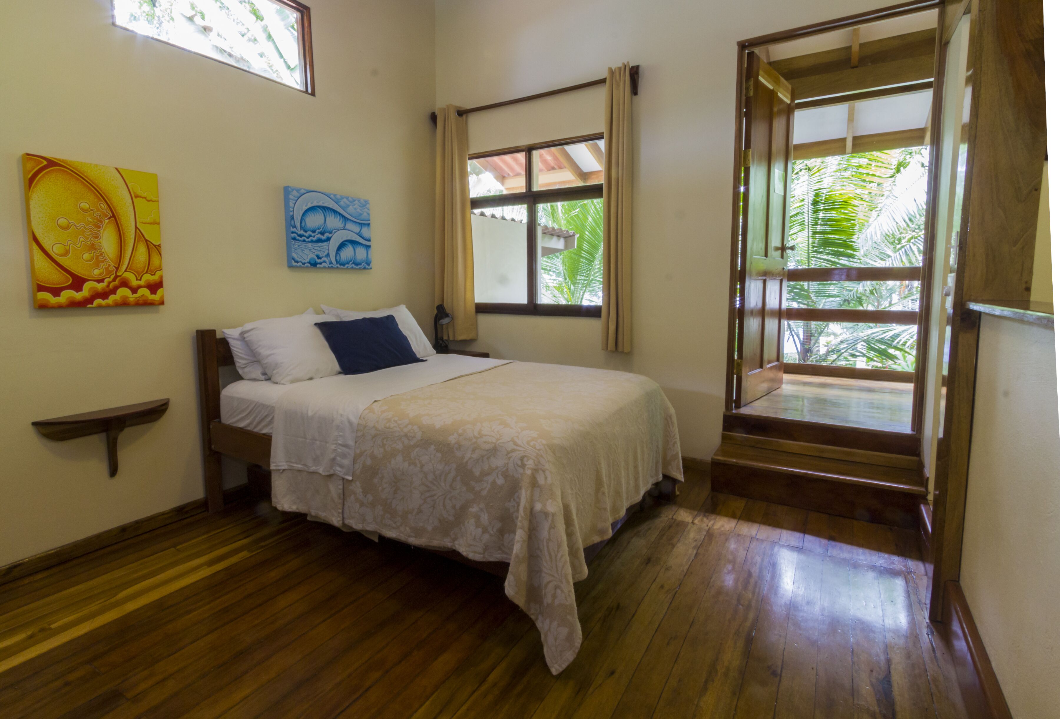 Une chambre à coucher avec un lit, une fenêtre avec des rideaux, un plancher en bois et deux tableaux accrochés au mur.