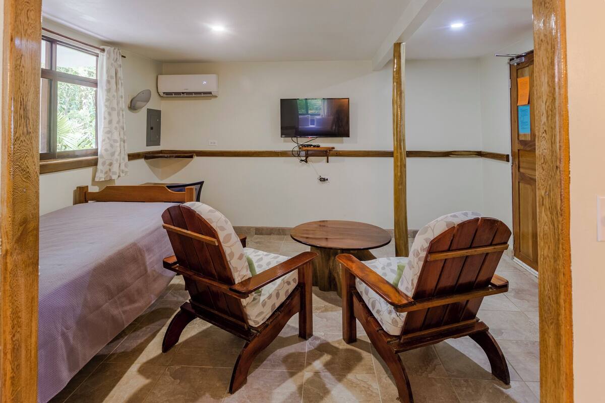 A room with a bed, two wooden chairs, a small round table, a TV mounted on the wall, and a window with curtains.