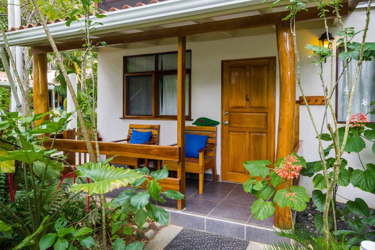 A wooden porch with benches and a door, surrounded by lush greenery.