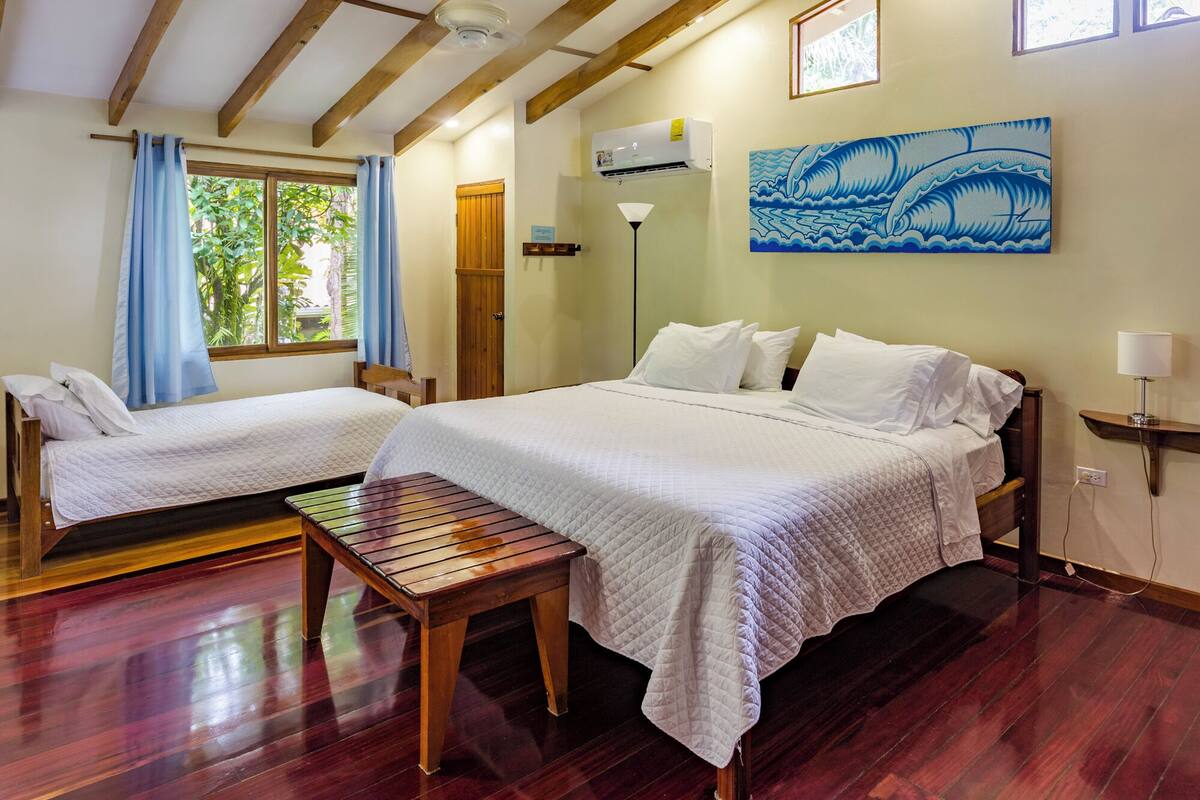 A room with two beds, a wooden bench, a ceiling fan, an air conditioning unit, and a painting of waves.