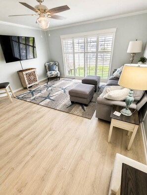 A living room with a ceiling fan, a flat-screen TV, a sofa, an ottoman, a coffee table, and a window with blinds.