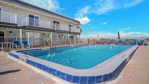 Et poolområde med en klar, blå pool, en bygning med altaner, og en indhegnet terrasse med liggestole.