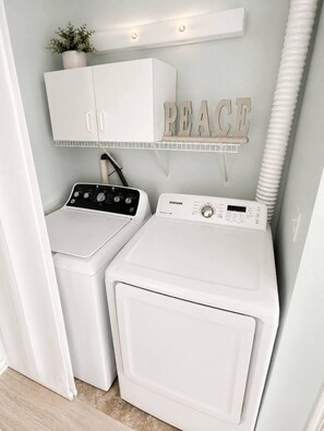 A laundry room with two white front-loading washing machines, a white cabinet above, and a shelf with the word 'PEACE' on it.
