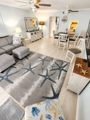 Una sala de estar con un sofá gris, un reposapiés y un tapete con estampado de estrella de mar. Hay una zona de comedor con sillas blancas y una mesa de madera.