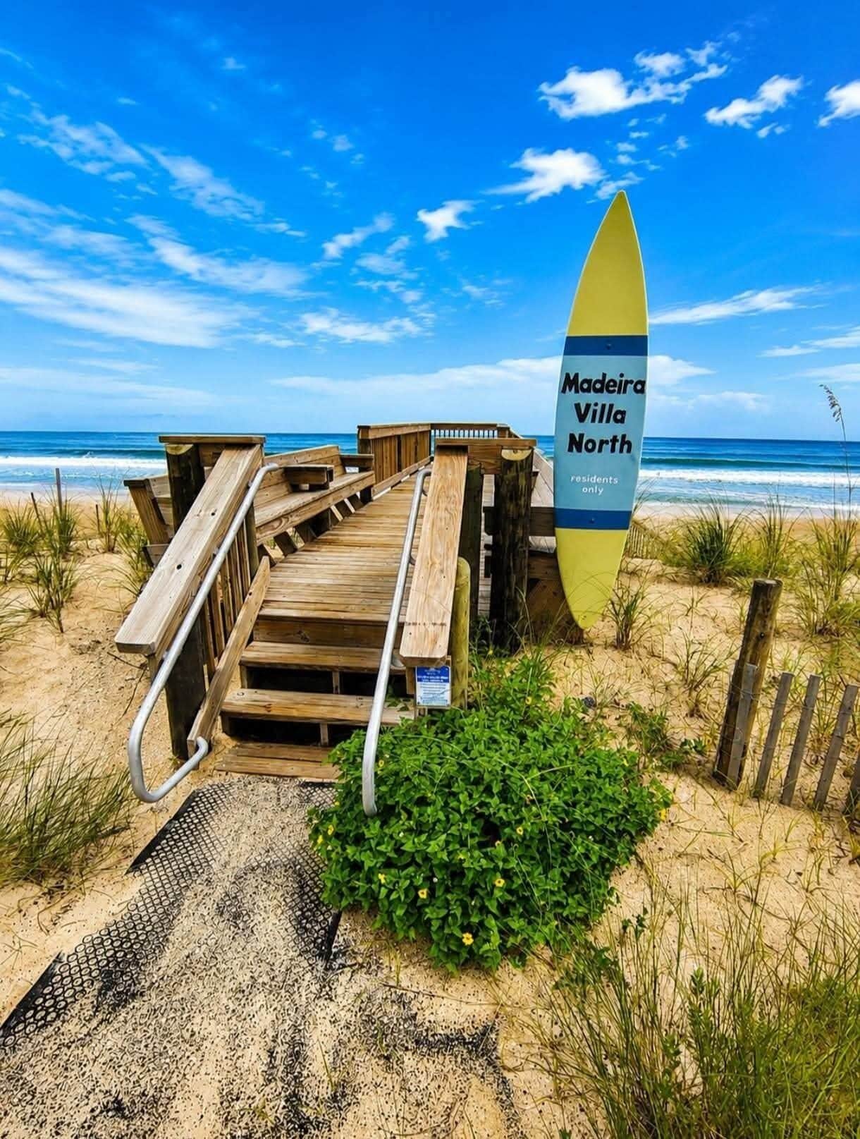 Een houten looppad naar een strand, met een geel surfboard en een bordje voor Madeira Villa North.