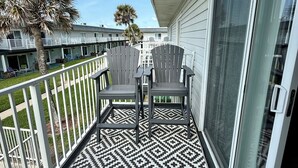 En balkon med Adirondack-stole, et mønstret tæppe og udsigt til omkringliggende bygninger.