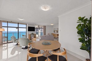 A modern living room with a large window overlooking the sea, a dining area with a round table, and a flat-screen TV mounted on the wall.