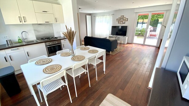 A modern kitchen with white cabinets, a dining area with a white table and chairs, and a living area with a dark sofa and a TV. - Superbe Maison - Biarritz/plage (Biarritz)