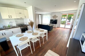 A modern kitchen with white cabinets, a dining area with a white table and chairs, and a living area with a dark sofa and a TV.
