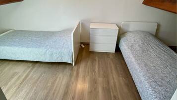 A small, compact room with two single beds, a white bedside table, and a wooden floor.