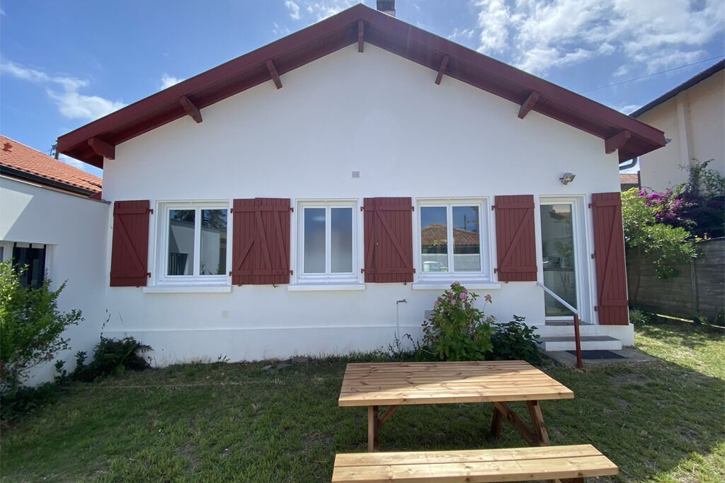 A single-story house with a white exterior, red shutters, and a wooden table in the front yard.